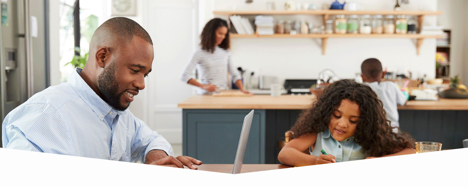 A young black family in the kitchen. Dad is working on his laptop. The daughter is coloring next to him at the table. Mother and son work on something at the kitchen island.